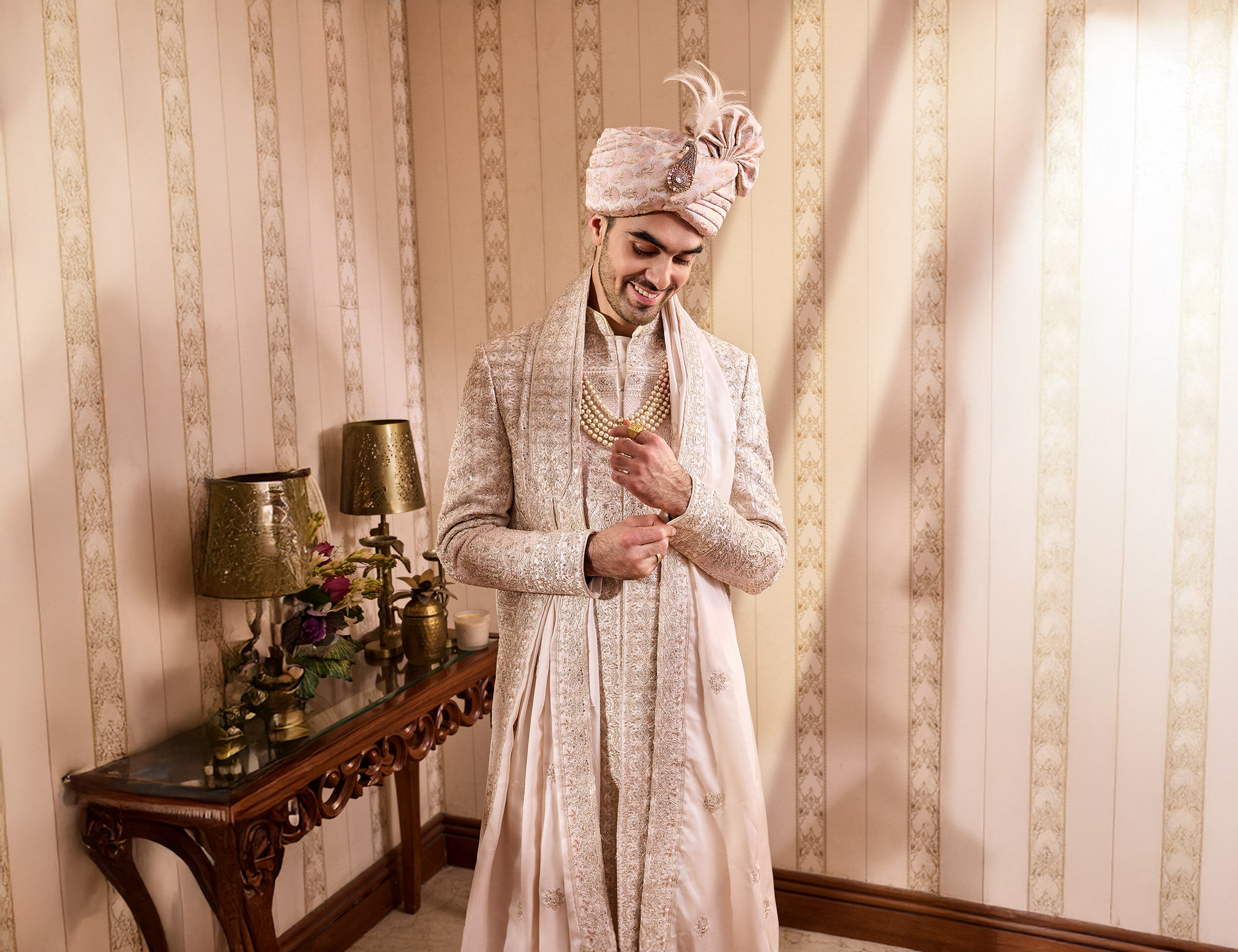 Groom in pastel embroidered sherwani with matching turban and layered pearl necklace, posing indoors before wedding